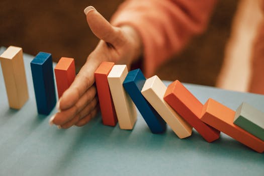 pexels-photo-12955678-12955678 A hand strategically stops falling blue and red domino blocks on a table.
